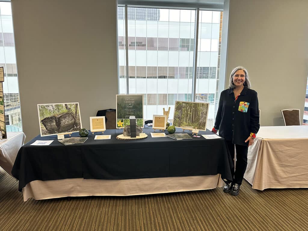 Katherine standing beside an exhibition table with various display items and informational placards inside a room with large windows.