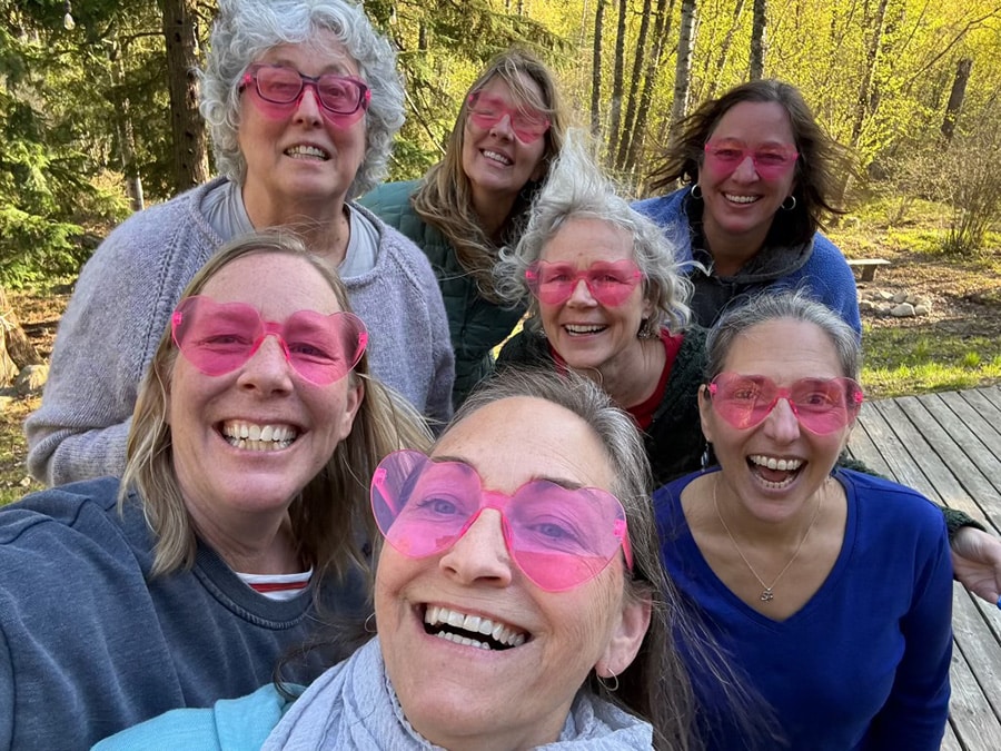 A group of seven smiling people, all wearing pink heart-shaped sunglasses, pose together outdoors in a forested area with green trees in the background.