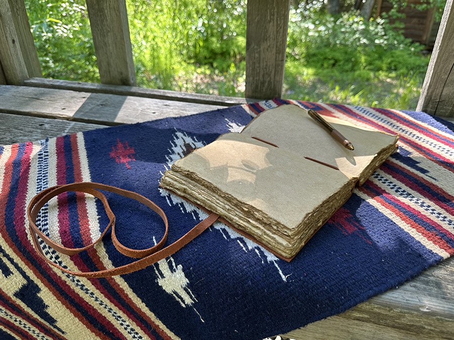 An open, weathered notebook with a leather strap lies on a patterned cloth on a wooden bench, accompanied by a pen. The background shows a lush, green outdoor area.