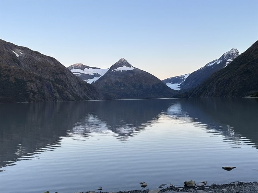 A serene lake reflects nearby mountains with snow-capped peaks under a clear sky, creating an image worthy of any media spotlight. Stones line the shore in the foreground.