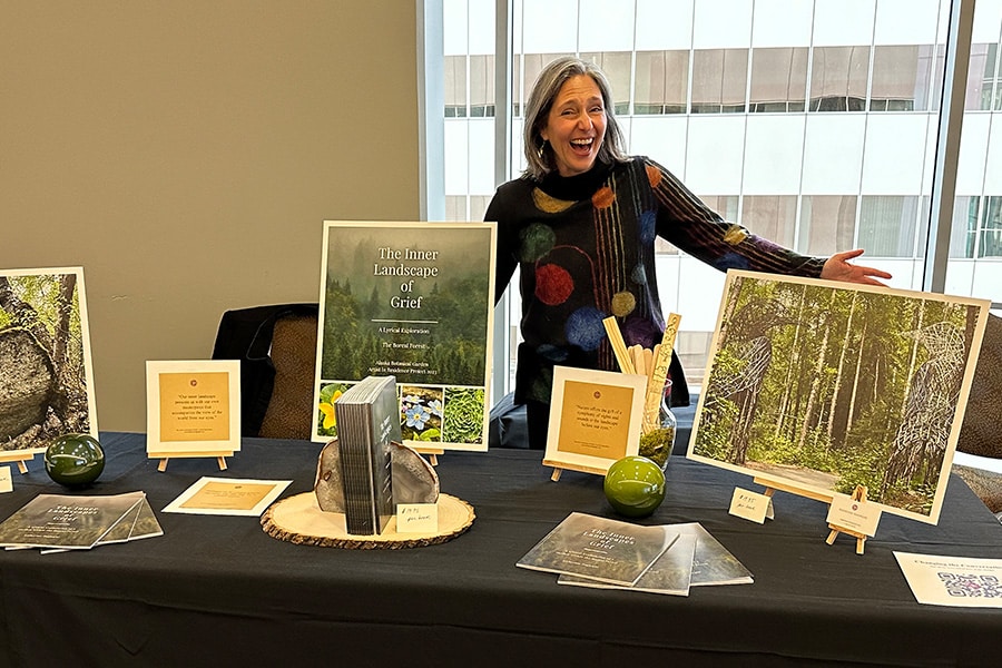 A smiling woman stands behind a table displaying books and photographs of nature, including a book titled "The Inner Landscape of Grief." The table is set up in front of a wall of windows.