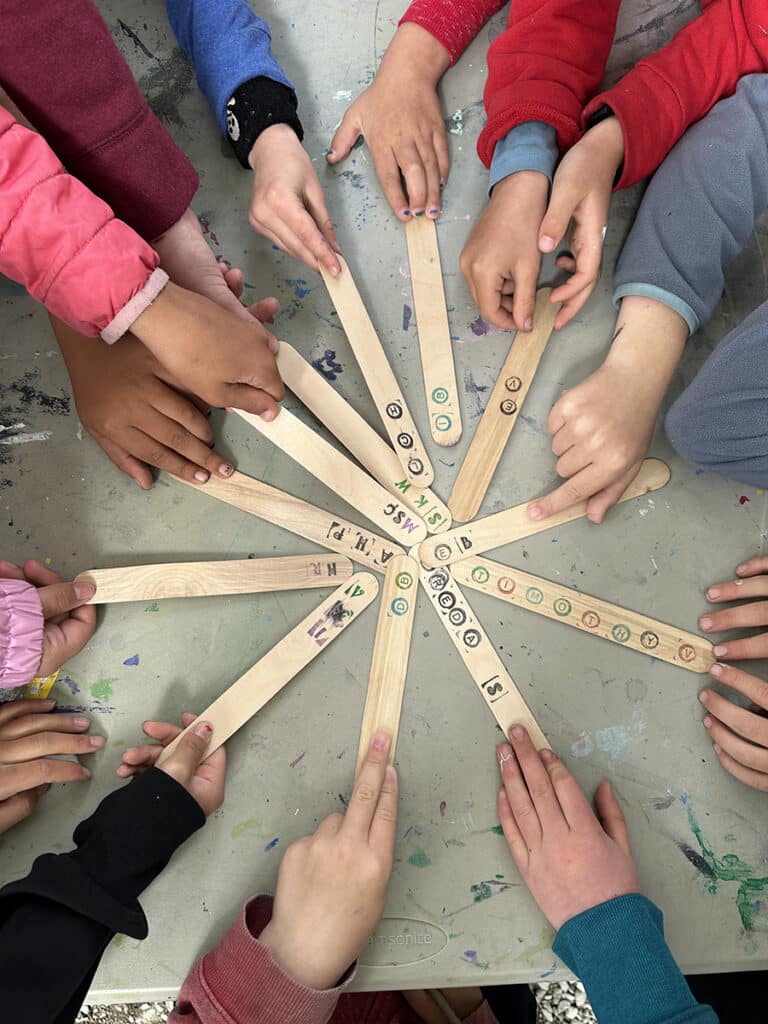 Children's hands are pointing at wooden sticks arranged in a star pattern on a table. The sticks are marked with various letters, numbers, and symbols.