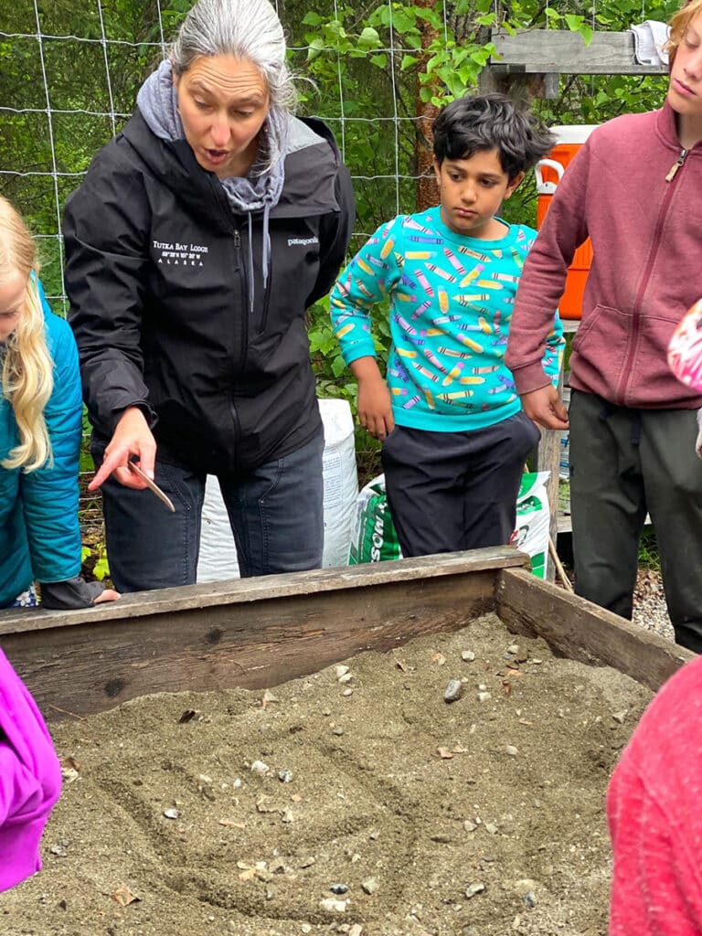 A woman points and explains something to a group of children gathered around a wooden sandbox filled with sand and small rocks, outdoors. Various gardening supplies are visible in the background.